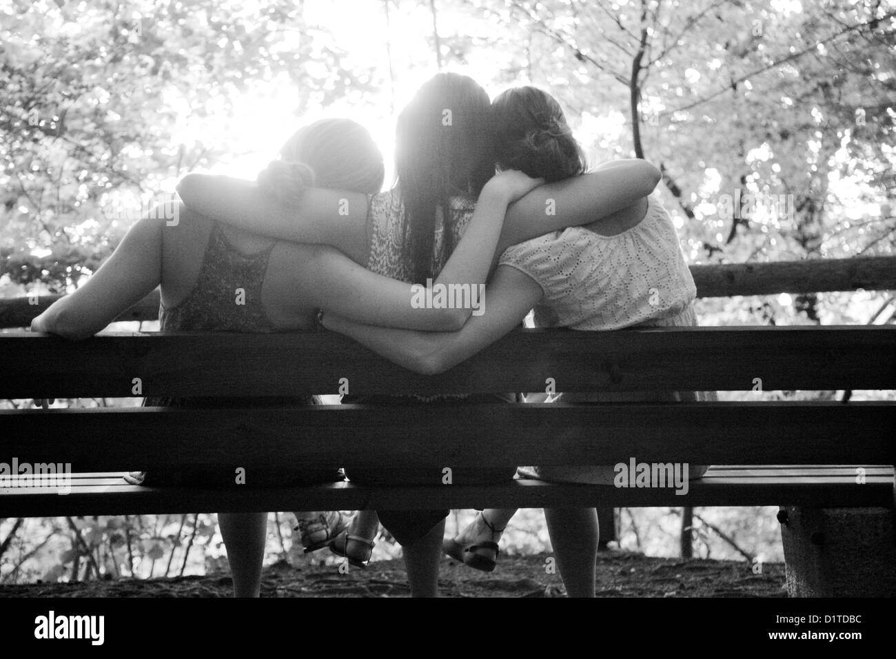 Eine Gruppe von drei Mädchen, mit dem Rücken zur Kamera, sitzen auf einer Bank, Arme Runde miteinander, mit Blick auf die Sonne und die Bäume. Stockfoto