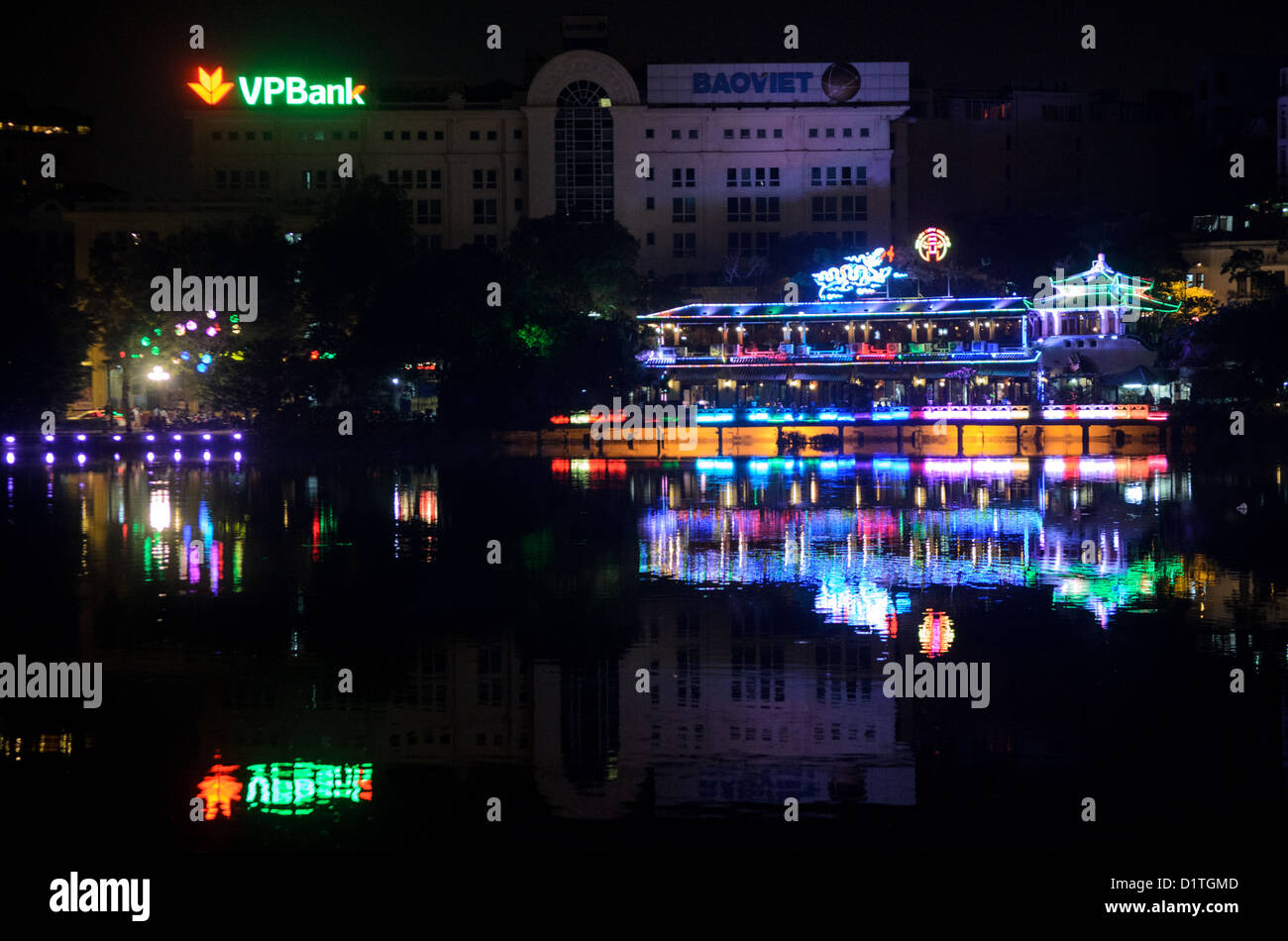 Hoan Kiem Lake Nachtansicht Hanoi Vietnam // HANOI, Vietnam — Stadtlichter von Gebäuden entlang der nordwestlichen Küste des Hoan Kiem Lake erzeugen Reflexionen auf der ruhigen Wasseroberfläche. Der historische See in der Altstadt von Hanoi ist ein zentrales Merkmal der nächtlichen Landschaft der Stadt. Die Uferpromenade verbindet Architektur aus der Kolonialzeit mit moderner Beleuchtung. Stockfoto