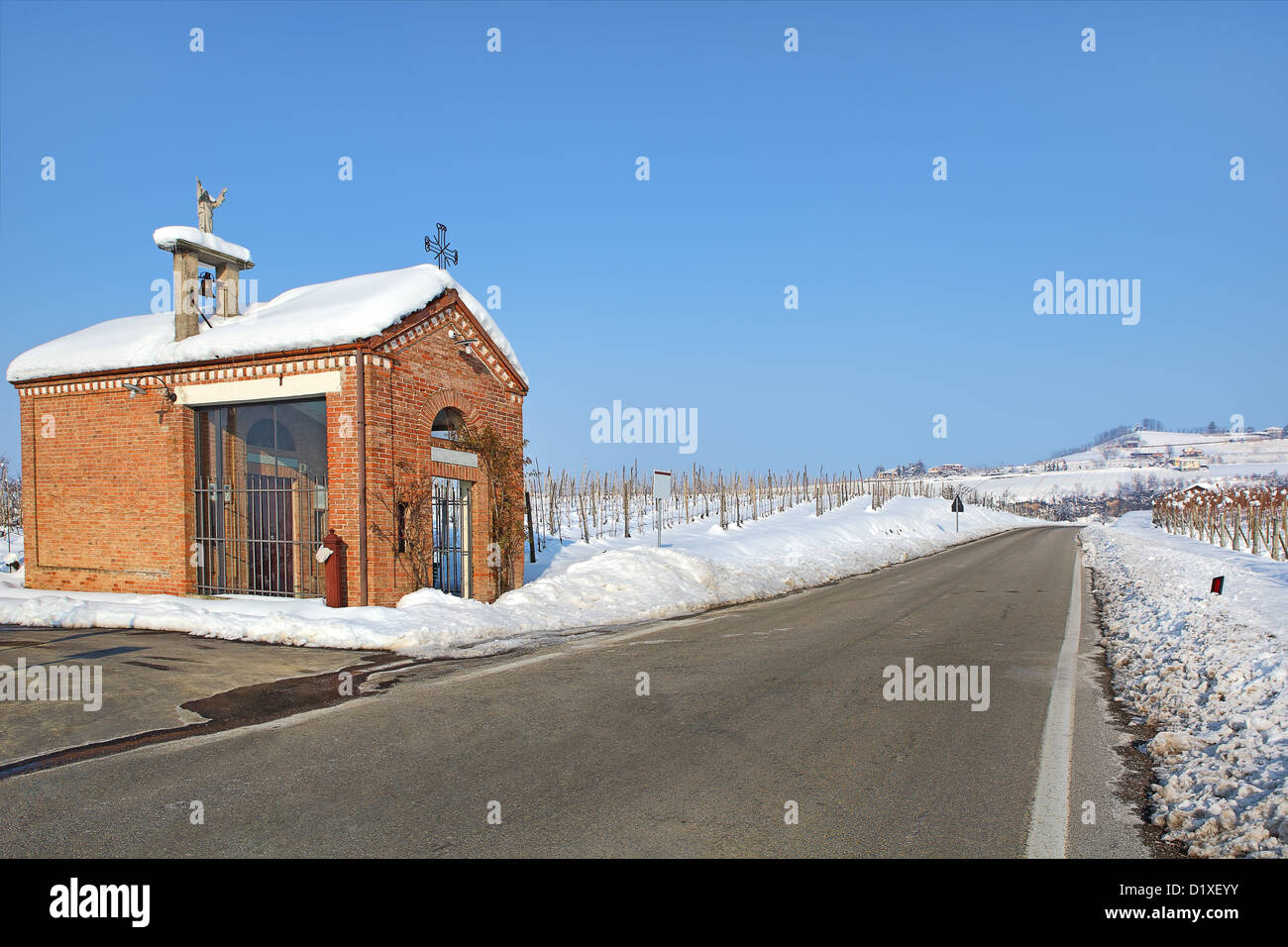 Kleine Rote Kapelle am Straßenrand zwischen Weinbergen, die unter dem weißen Schnee unter klaren, blauen Winterhimmel im Piemont, Norditalien. Stockfoto