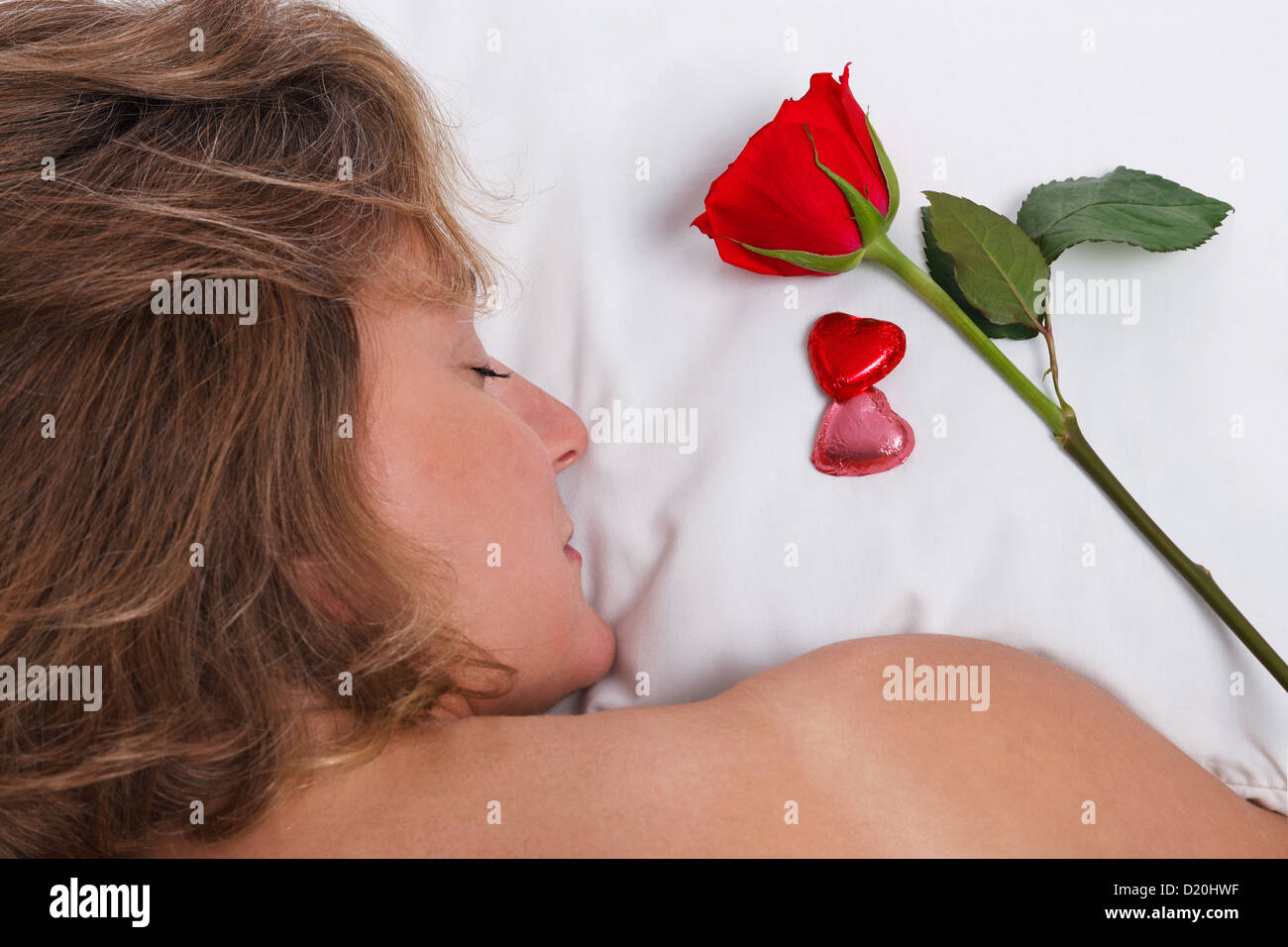 Eine Frau schläft, während für den Valentinstag eine einzelne rote Rose und herzförmige Pralinen auf ihrem Kissen geblieben sind. Stockfoto