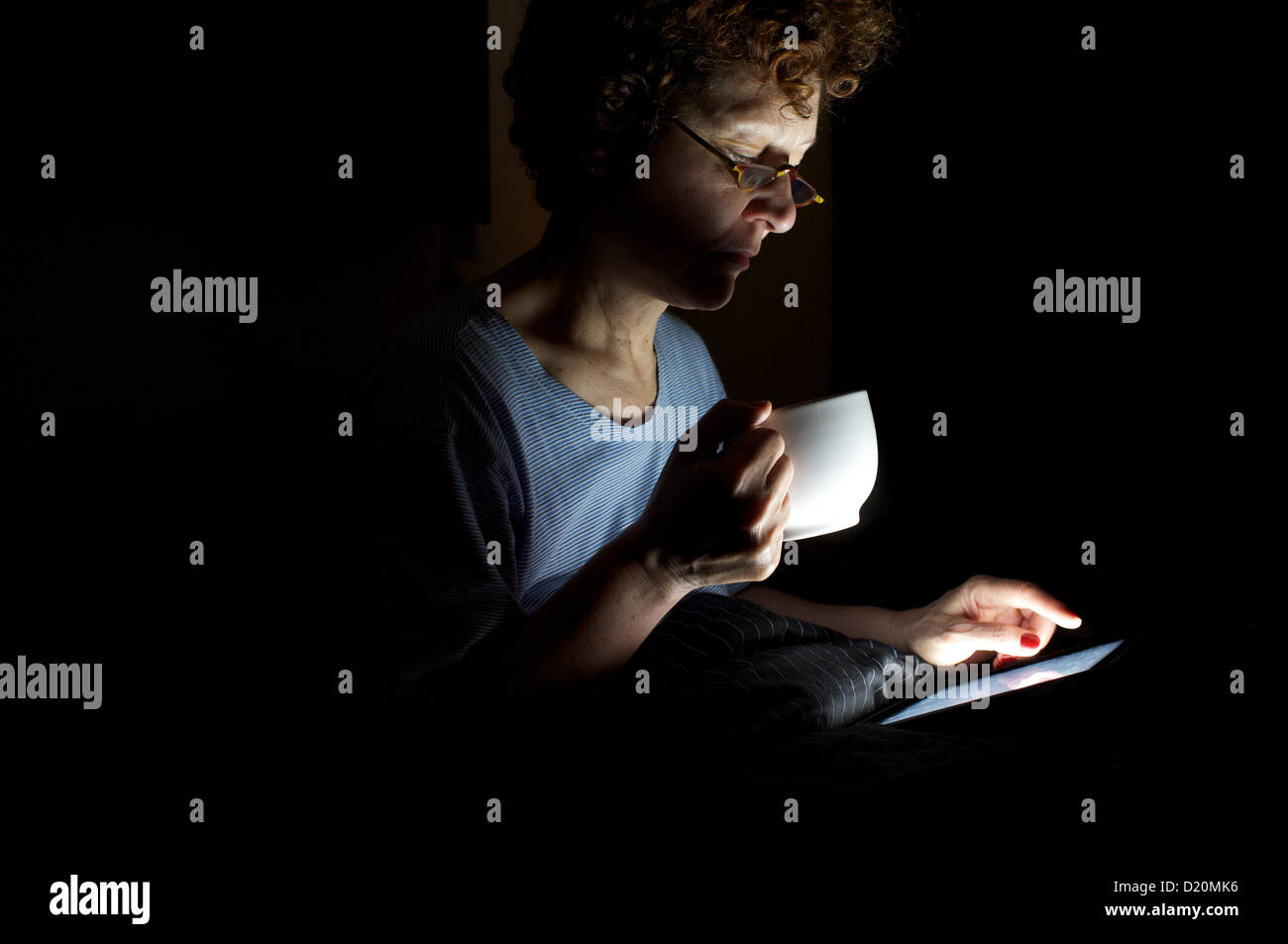 Frau trinkt eine Tasse Kaffee beim Lesen eines Buches auf einen e-Reader im Bett sitzend Stockfoto