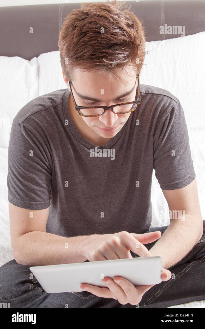 Junger Mann mit Brille auf den Bildschirm von seiner digital-Tablette, sitzt auf seinem Bett. Stockfoto
