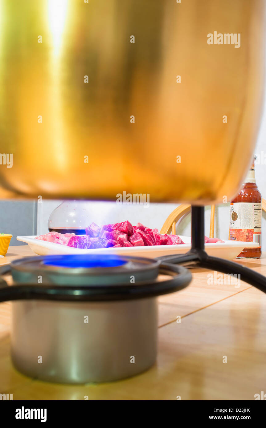 Fleisch-Fondue-Topf mit Öl erhitzen über offener Flamme aus der Dose Kochen von Brennstoff, Teller mit rohem Fleisch im Hintergrund gefüllt Stockfoto