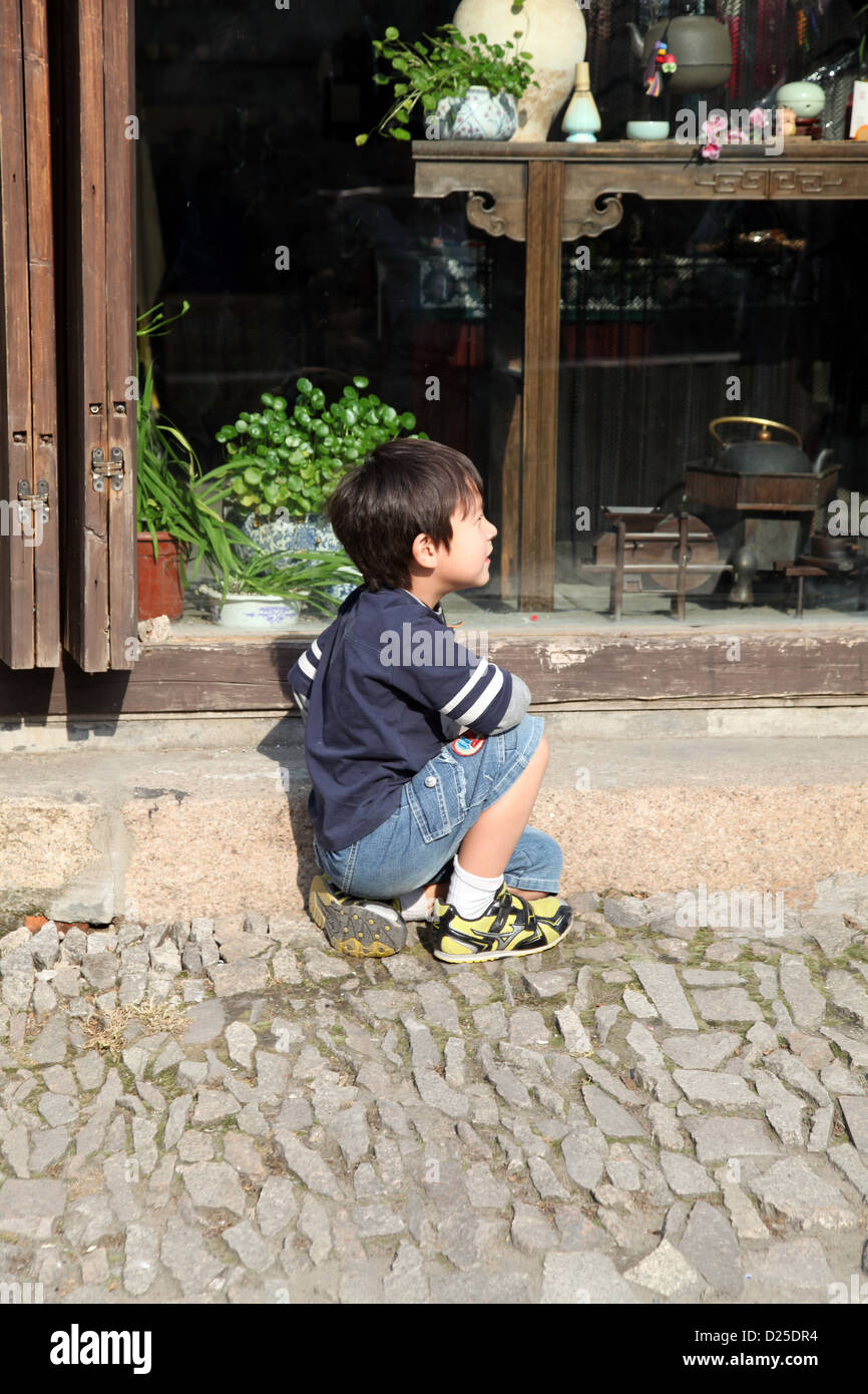 Es ist ein Foto eines chinesischen jungen, die sitzt auf dem Boden vor einem Fenster ein Souvenir-Shop. Er schaut nach rechts. Stockfoto