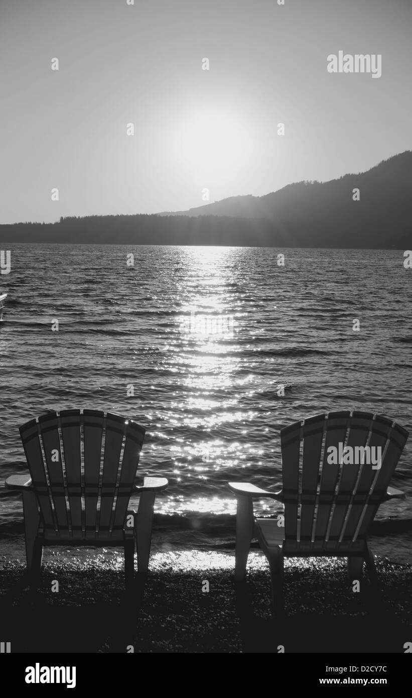 Graustufen-Bild 2 Adirondack Stühle auf einem Steinstrand mit die untergehende Sonne in der Ferne. Stockfoto