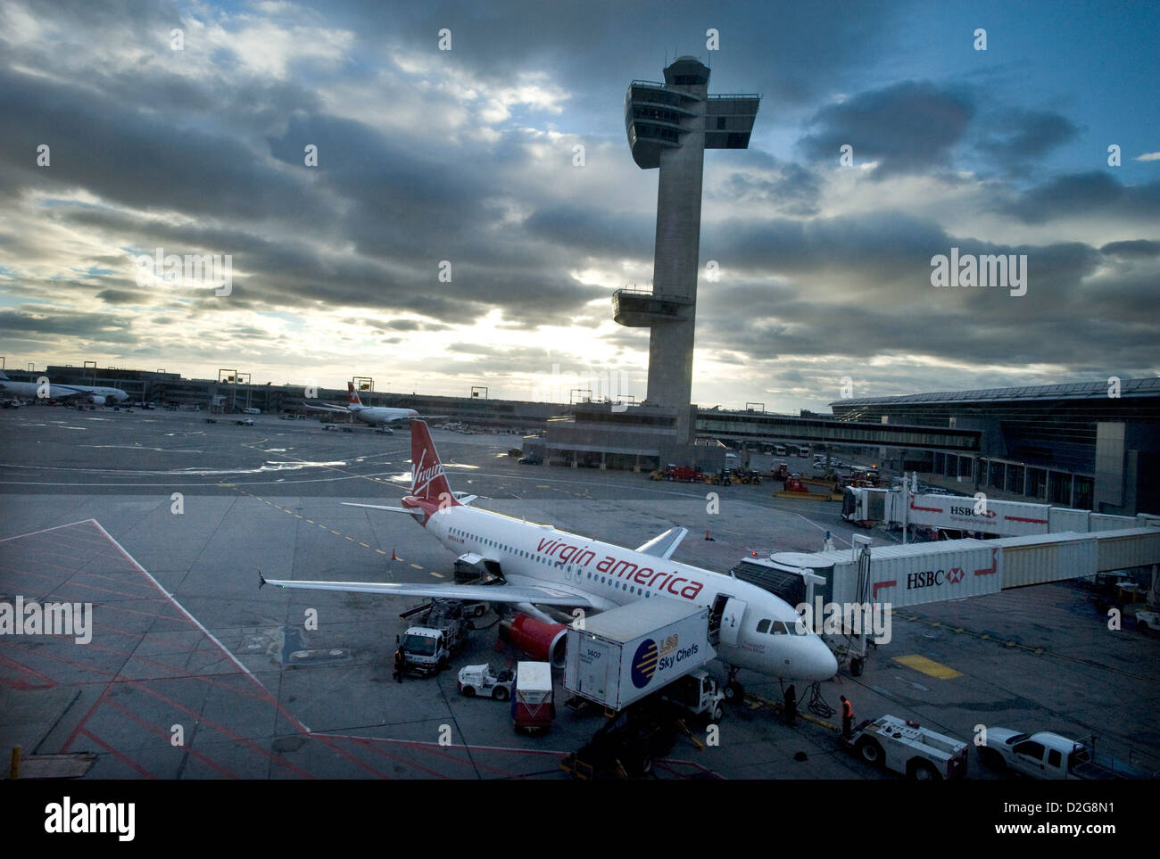 Eine Jungfrau Amerika Airbus A320 Jet steht Flugzeuge vor dem Kontrollturm in der Dämmerung, wie es geladen mit Essen ist und von LSG Sky Chefs am John F. Kennedy Airport, JFK Airport, New York, USA beliefert Stockfoto