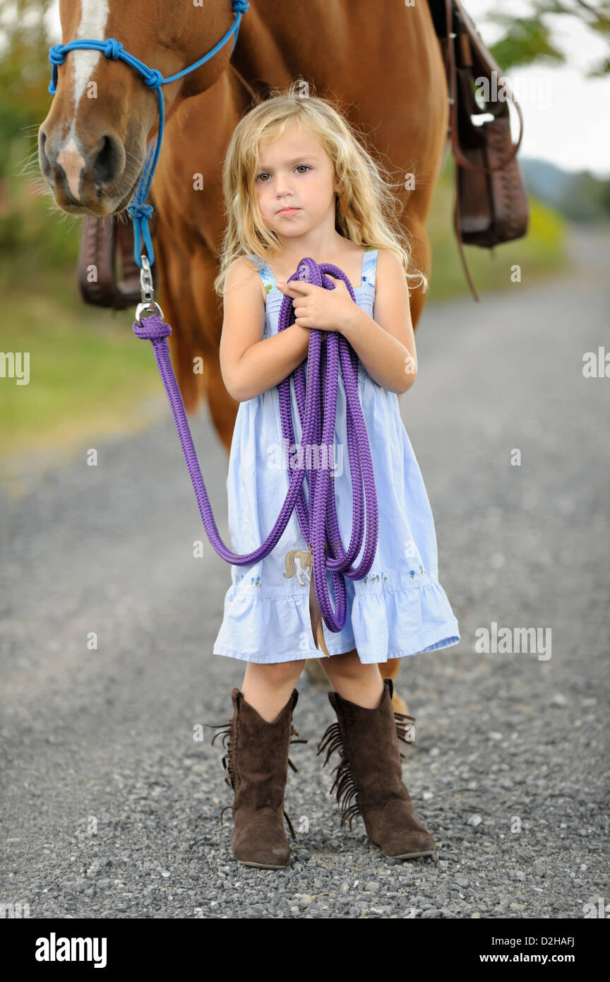 Kleine blonde Mädchen posiert mit ihren großen Pferd auf einen Feldweg, zuversichtlich Pose mit stetigen Blick, Stiefel blaue Sonne Kleid und hoch. Stockfoto