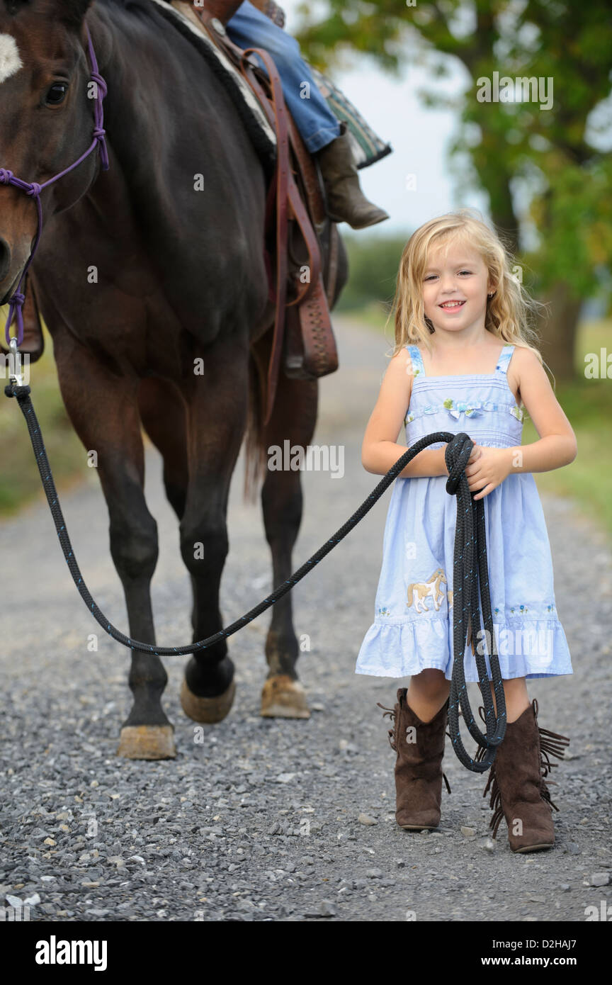 Kleine blonde Cowgirl führt ihr großes Pferd auf einen Feldweg, Stiefel Lächeln auf den Lippen und tragen blaue Kleid und westlichen. Stockfoto