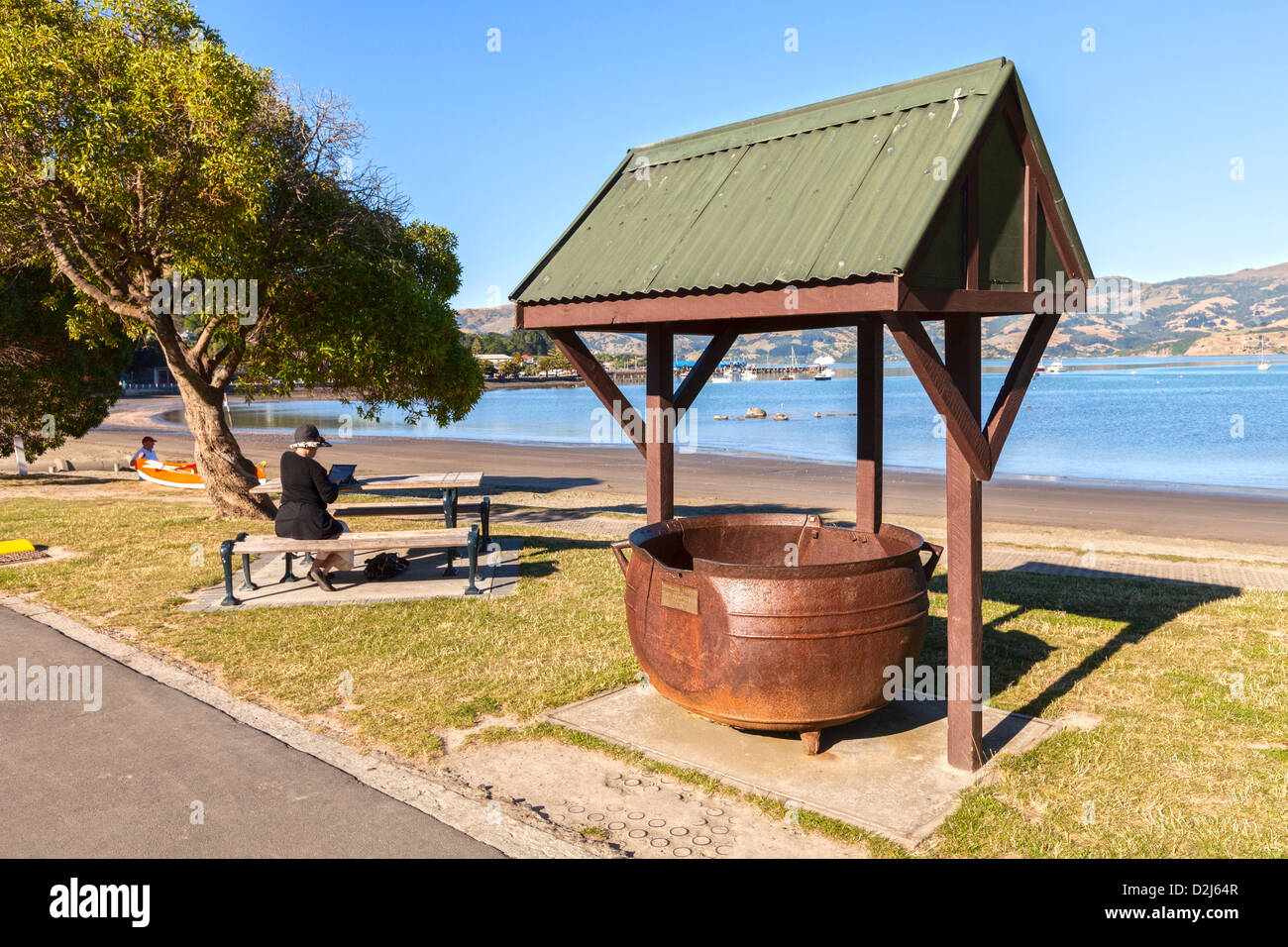 Ein Versuch Topf, Relikt der Akaroa Tage als eine Walfangstation, am Ufer des Hafens. Frau sitzt auf der Bank Usig iPad. Stockfoto