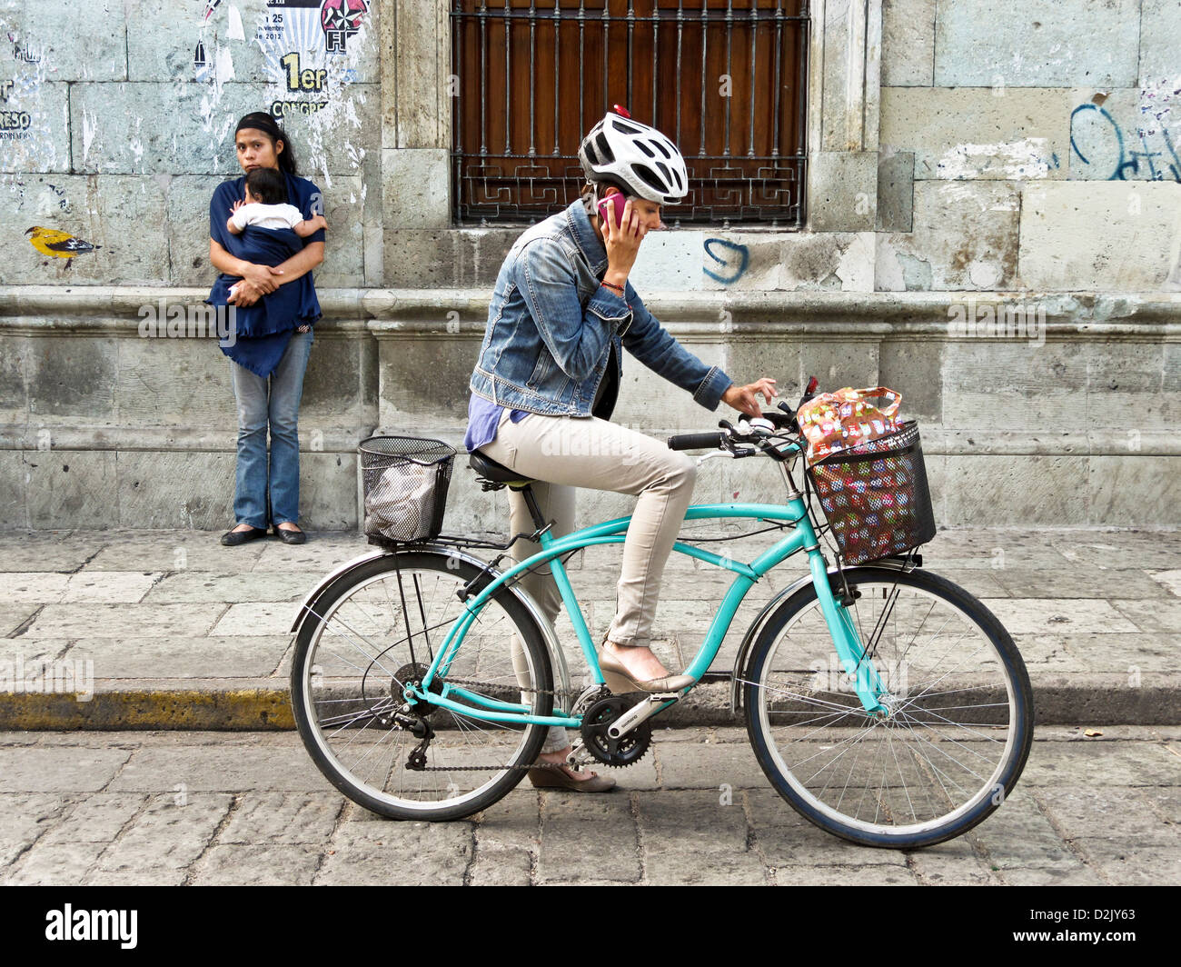 moderne junge Mexikanerin auf dem Fahrrad mit Handy Sturzhelm & indigene indische Teenager mit Baby auf Bürgersteig Oaxaca Stockfoto