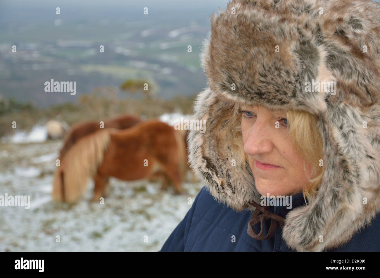 Reife Frau in Pelzmütze wegsehen von Kamera mit Schnee und Ponys im Hintergrund Stockfoto