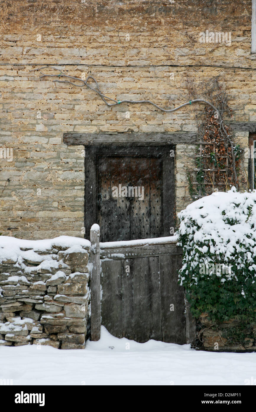 Cotswold reetgedeckten Landhaus aus Stein mit hölzernen Eingangstür, Gartenmauer und Holztor. Schnee fällt und liegt auf dem Boden. Stockfoto