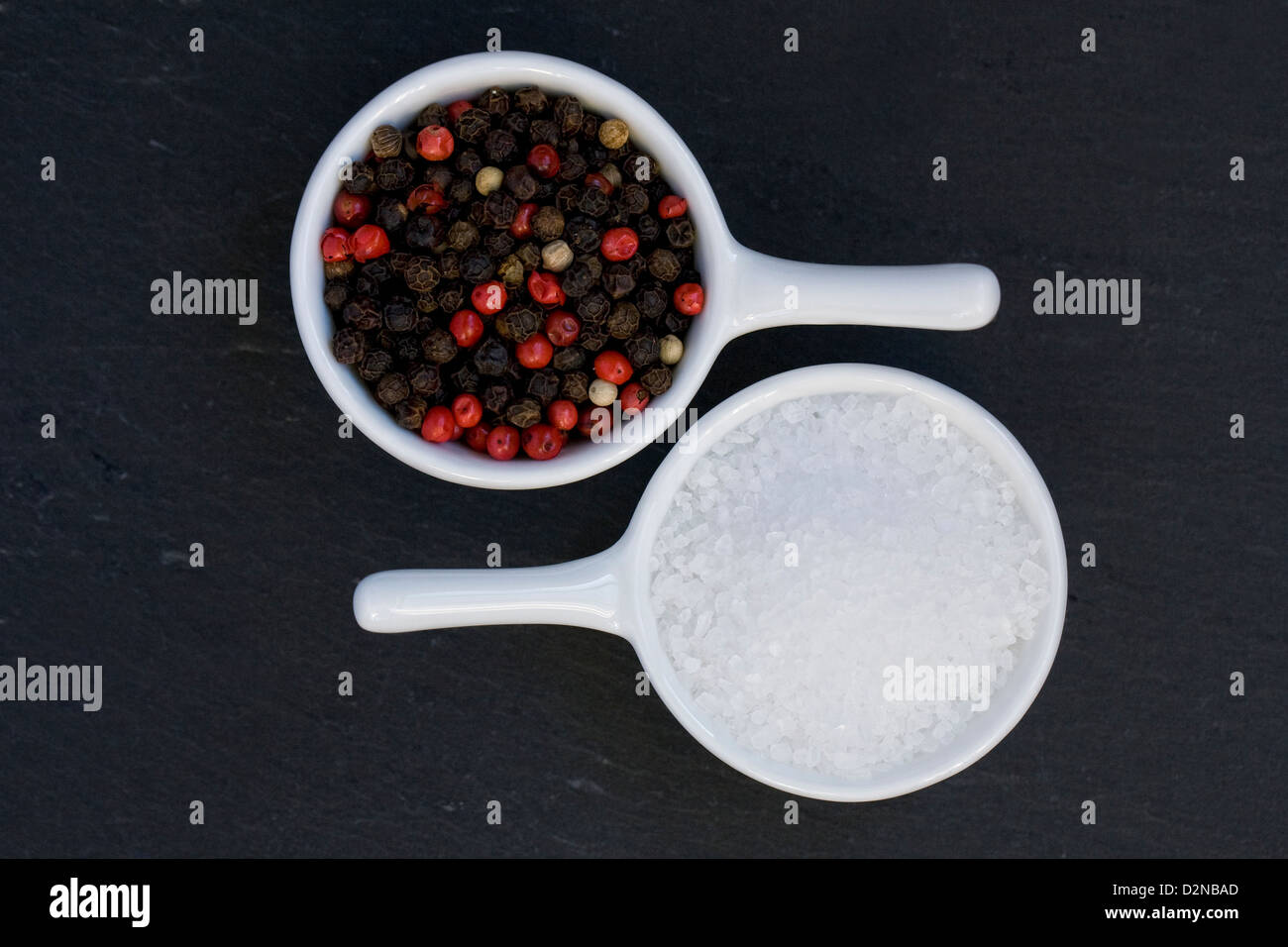 Zwei kleine weiße Gerichte mit Pfefferkörnern und Seasalt vor einem schwarzen Hintergrund. Stockfoto