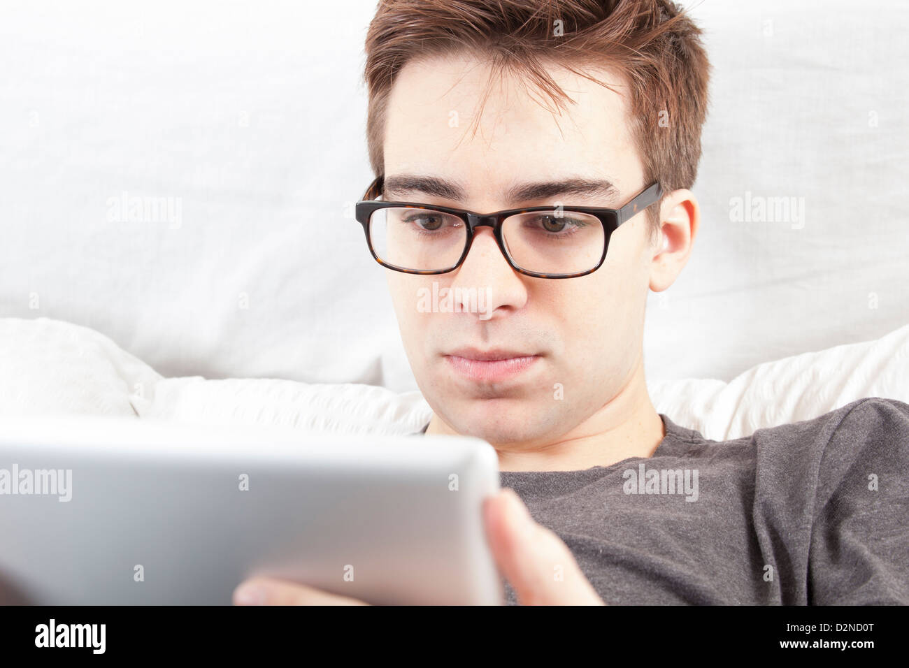 Junger Mann mit Brille auf den Bildschirm von seiner digital-Tablette, sitzt auf seinem Bett. Stockfoto