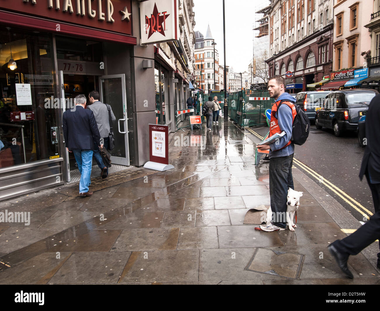 ein Mann verkauft Big Issue Papers im Regen mit seinem Hund auf einer Londoner Straße vor einem Restaurant der Stockfoto