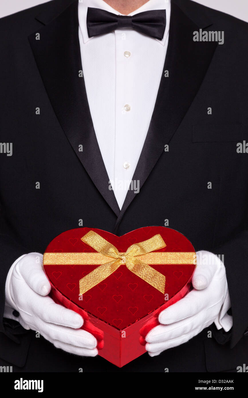Mann trägt schwarze Krawatte und weiße Handschuhe halten eine rote herzförmige Schachtel Pralinen. Stockfoto