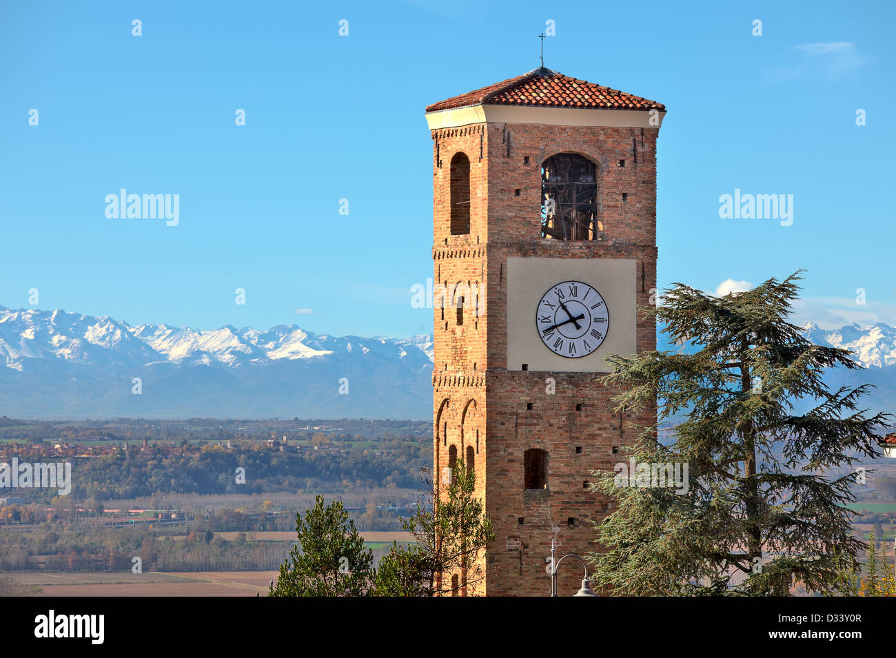 Blick auf alte gemauerte Glockenturm mit großer Uhr und Berge mit schneebedeckten Gipfeln im Hintergrund unter blauem Himmel in Piemont, Italien. Stockfoto
