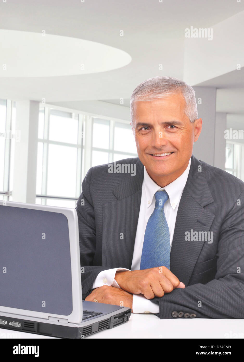 Im mittleren Alter Geschäftsmann sitzt an seinem Schreibtisch in einem modernen Büro mit Laptop. Mann lächelt in die Kamera. Hochformat. Stockfoto