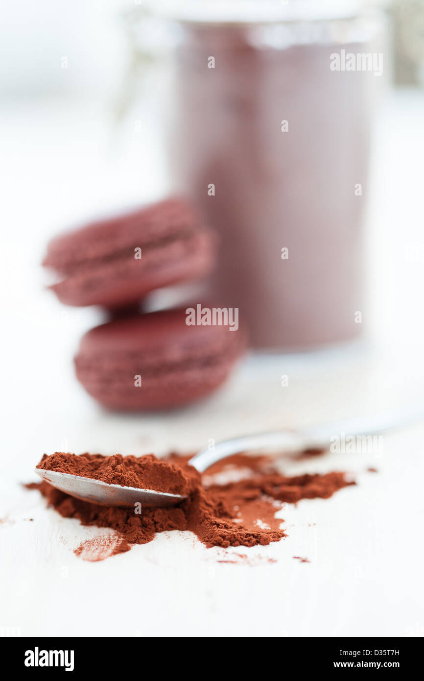 Nahaufnahme von Kakaopulver und Löffel mit zwei Makronen und Glas im Hintergrund Stockfoto
