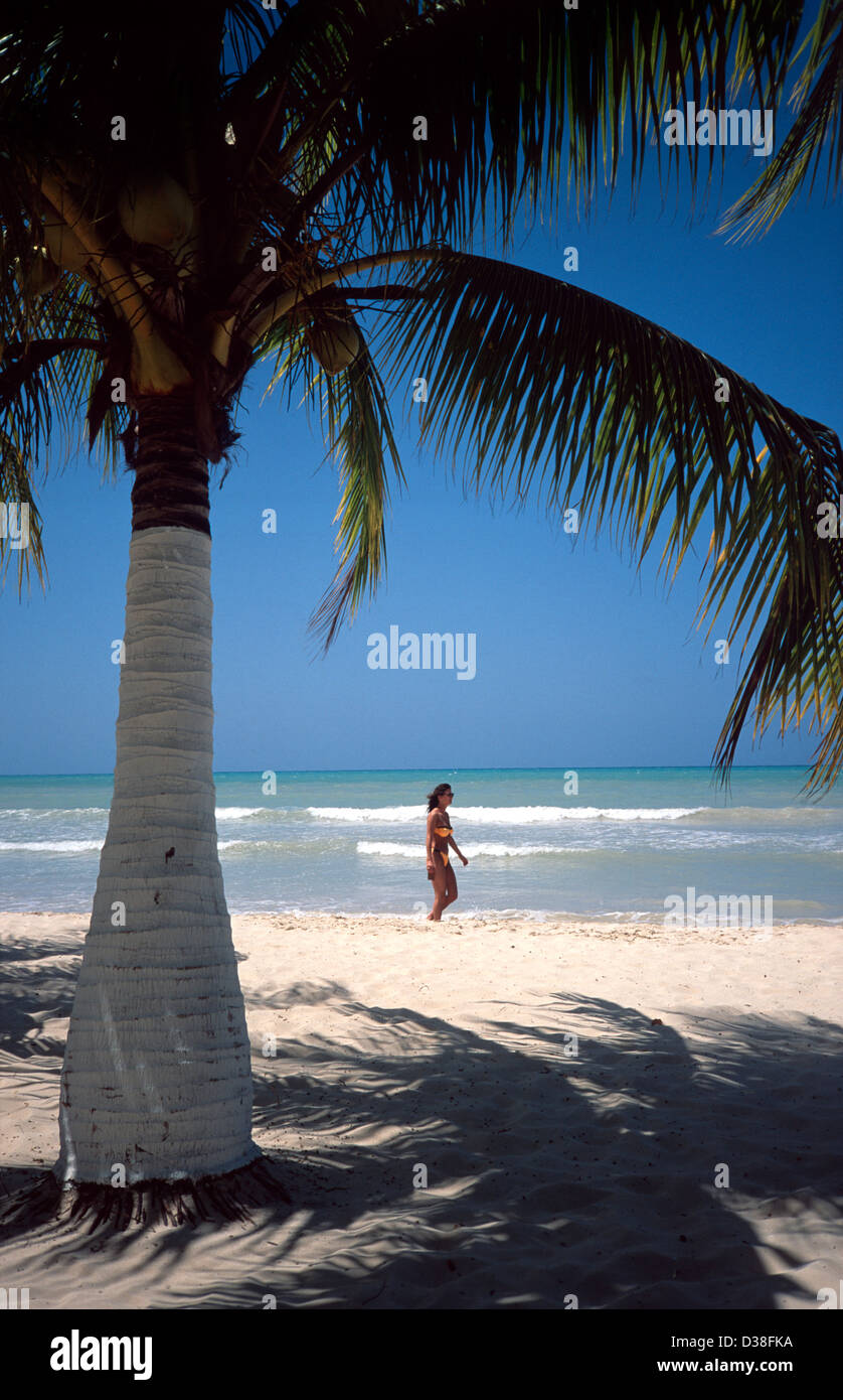 Frau in einem gelben Bikini, die am Strand von Negril spazieren geht, eingerahmt von einer Palme. Negril, Jamaika Stockfoto