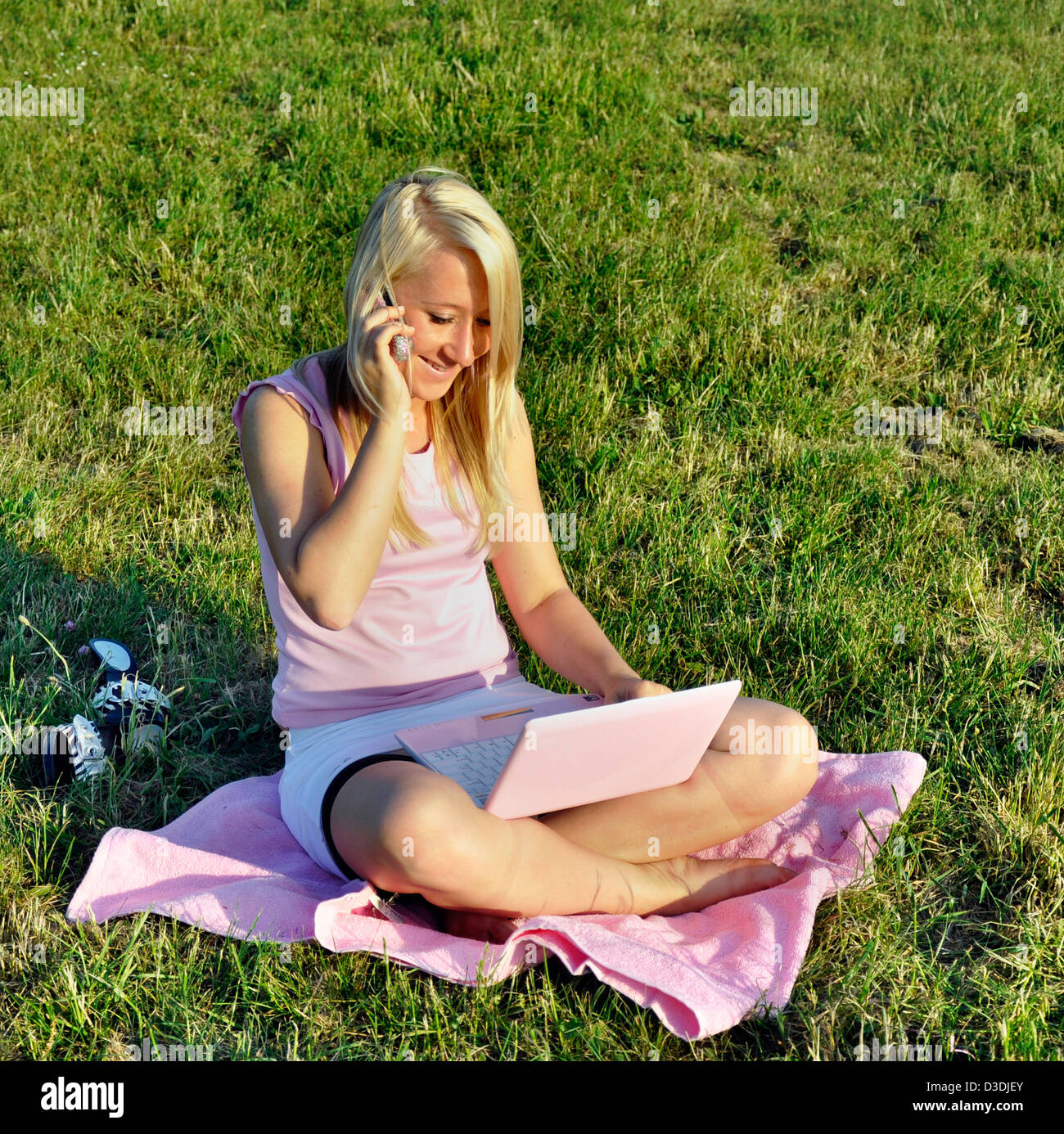 Ein Lächeln auf den Lippen entspannt Mädchen sitzt im Schneidersitz auf dem Rasen auf rosa Handtuch mit rosa Netbook und im Gespräch mit dem Mobiltelefon genutzt Stockfoto