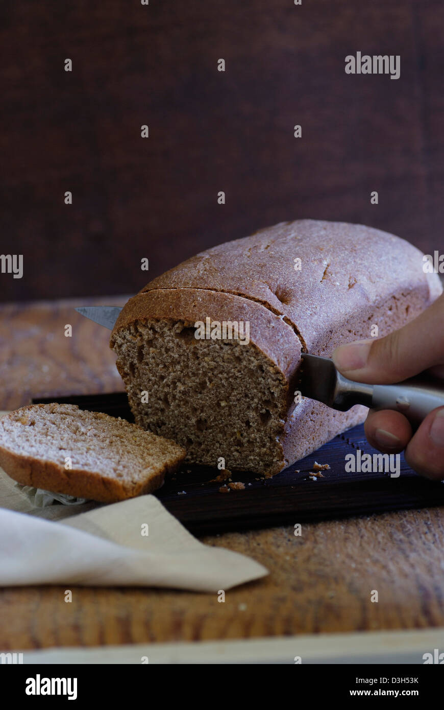 Artisan ganze Weizenbrot Brotlaib, Schneiden von Holz Brett Brotscheibe Messer von Hand Schneiden von Brot, Brot, weiße Stoffserviette Stockfoto