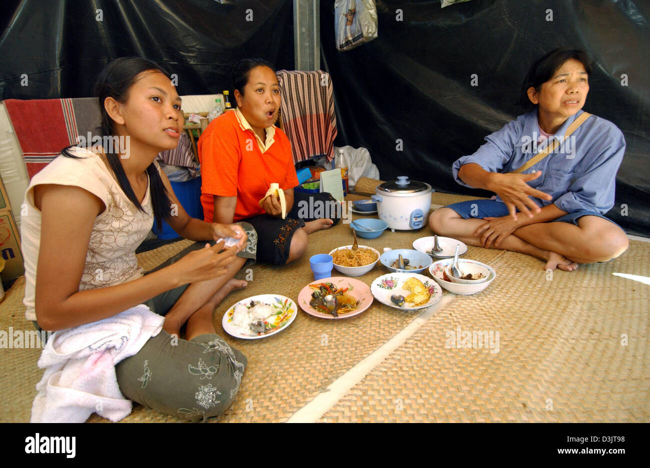 (Dpa) - sitzt, Chutipron Janakhon (L) zusammen mit ihrer Mutter Vasunan (C) und einem Freund in den Schatten einer Wellblech-Hütte während dem Mittagessen in einem Flüchtlingslager in der Nähe von Khao Lak, Thailand, 13. Januar 2005. Das Haus der Familie im Dorf Bang Neang wurde während der Flutkatastrophe vollständig zerstört. Der Vater und der Ehemann werden noch vermisst. Die Familie blickt auf den Bereich c Stockfoto