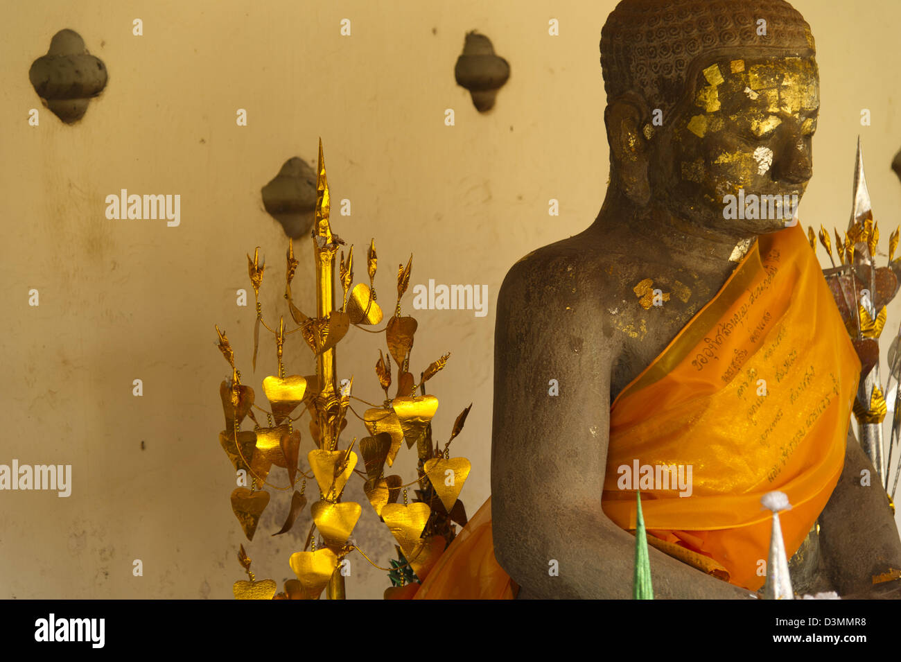 Eine Statue von Buddha drapiert in einer Safran Schärpe und Flecken von Blattgold sitzt neben einem hellen gold Angebot Baum bedeckt. Stockfoto