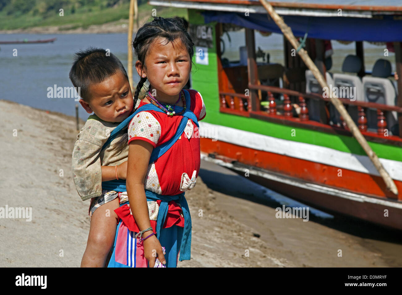 Mädchen mit Baby auf Rücken vor hölzernen Longtail-Boot entlang des Mekong in Laos, Südostasien Stockfoto