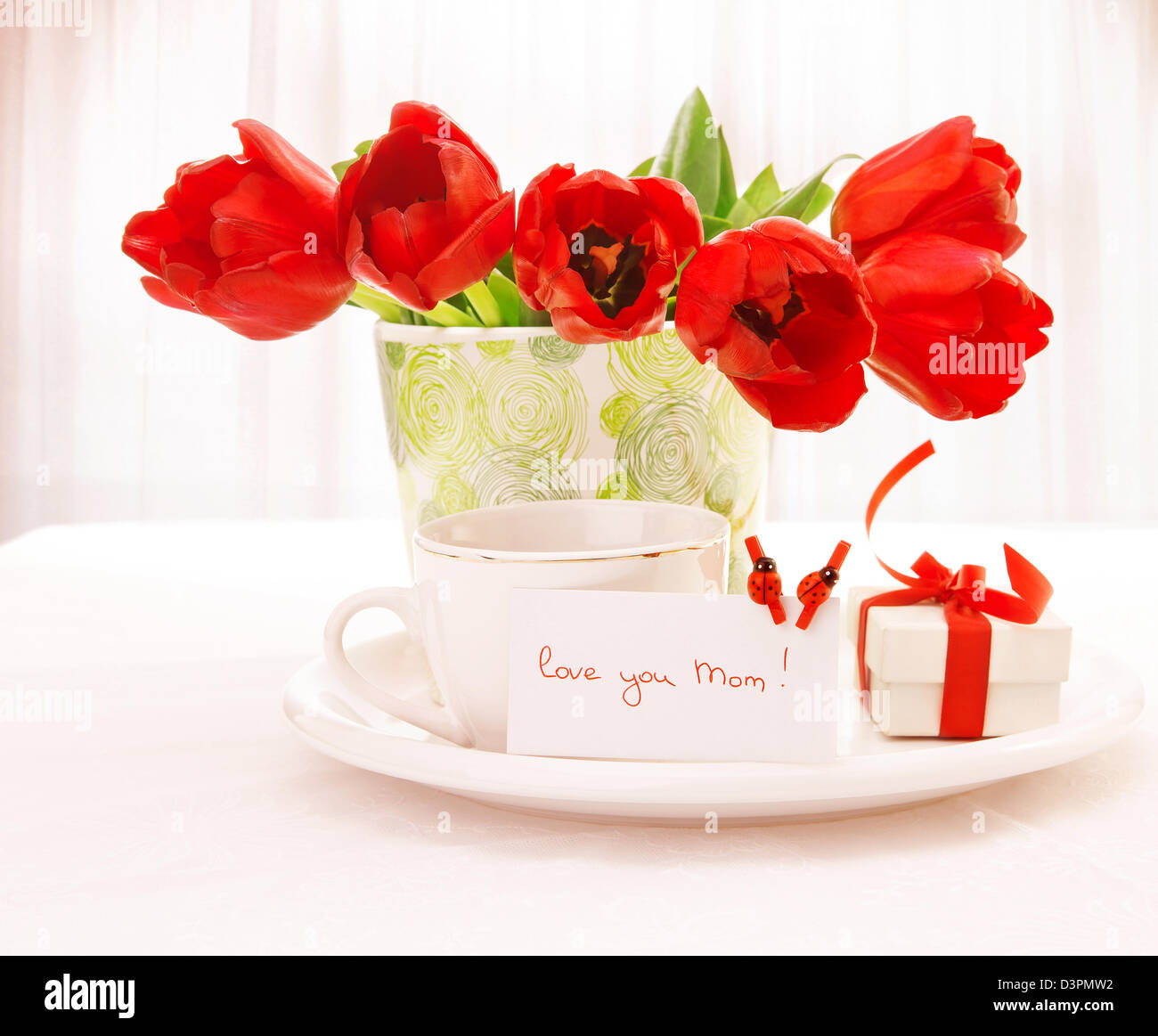 Foto von frische rote Tulpe Blumen in schönen Vase auf Tisch, Tasse Tee, kleinen weißen Geschenkbox mit Schleife, festliche Postkarte Stockfoto