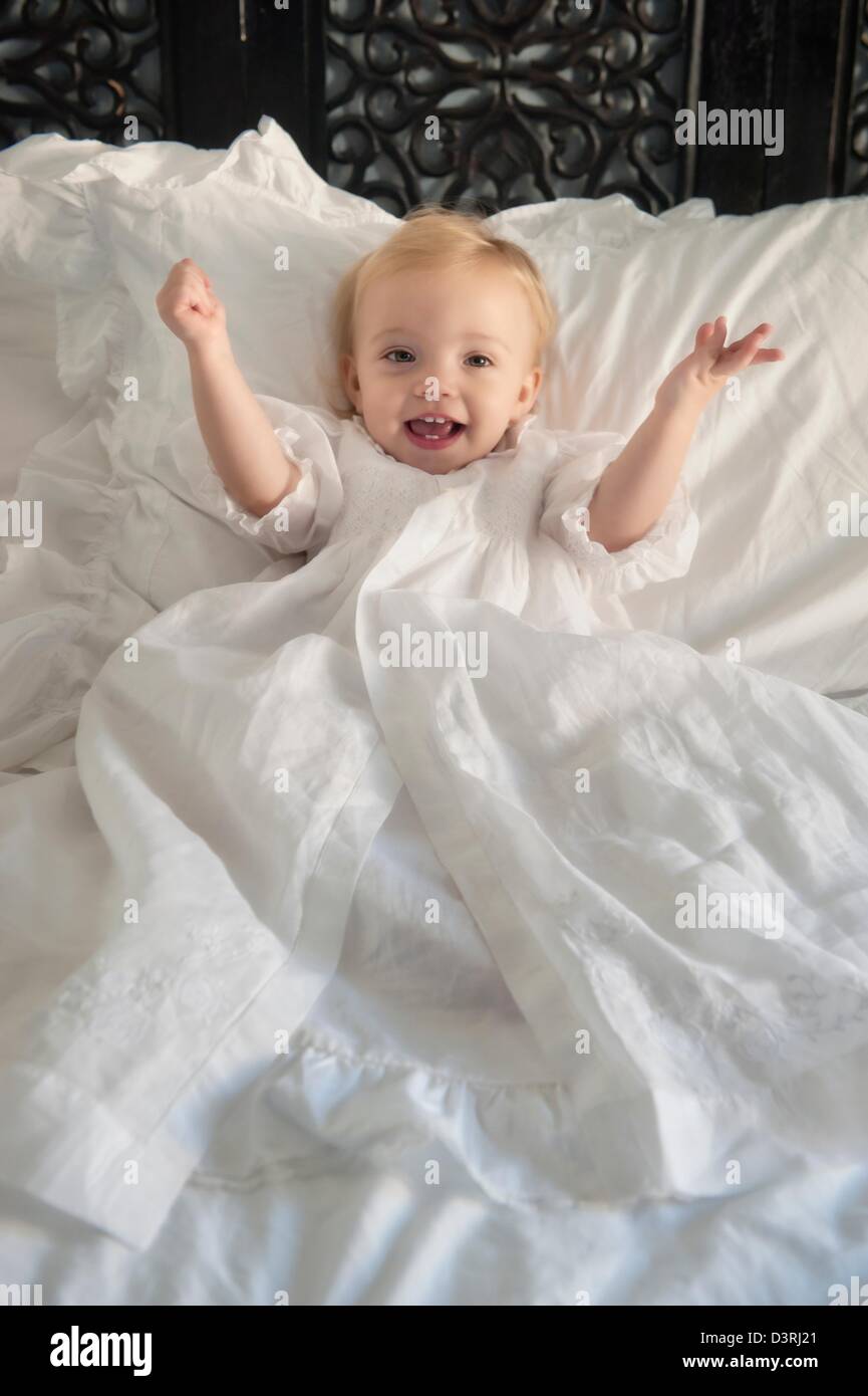 Glücklich ein 1 Jahr alt Baby Mädchen im Bett mit Armen liegend tragen weiße langes Kleid Kleid Taufe ausgestreckt Stockfoto