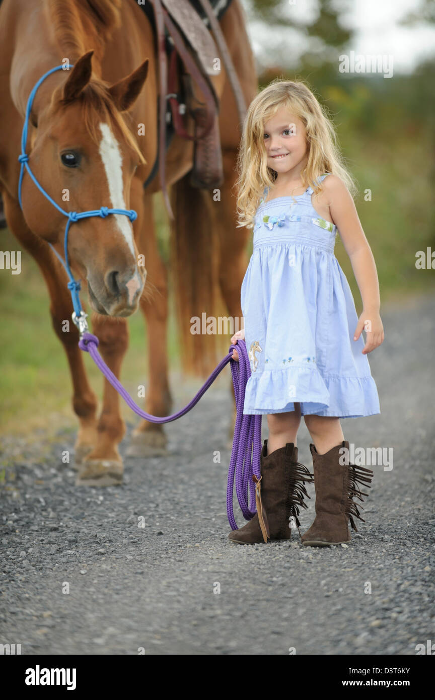 Kleine blonde Mädchen ihr großes Pferd auf einen Feldweg, lächelnd und glücklich 3 jährige trägt blaue Kleid und westlichen bo Stockfoto