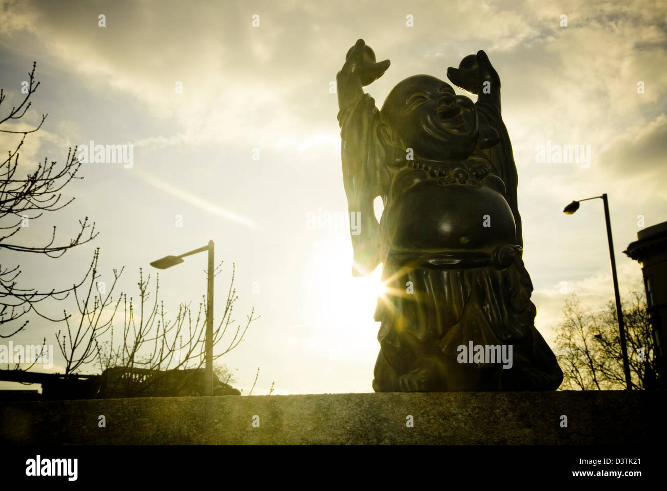 Buddha mit den Armen in der Luft sitzen auf einem Felsvorsprung mit goldenen Sonne von hinten kommende und Straße Laternenmasten im Hintergrund Stockfoto