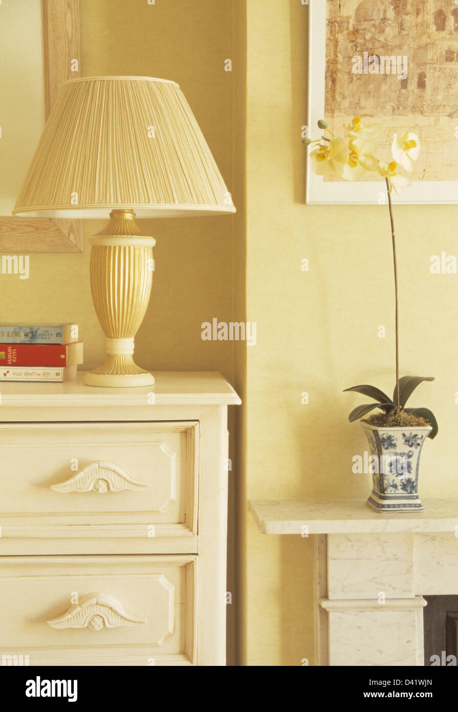 Blasse gelbe Schlafzimmer mit Creme Lampe mit plissierten Schirm auf der Kommode neben Kamin mit weiße Orchidee im Topf Stockfoto