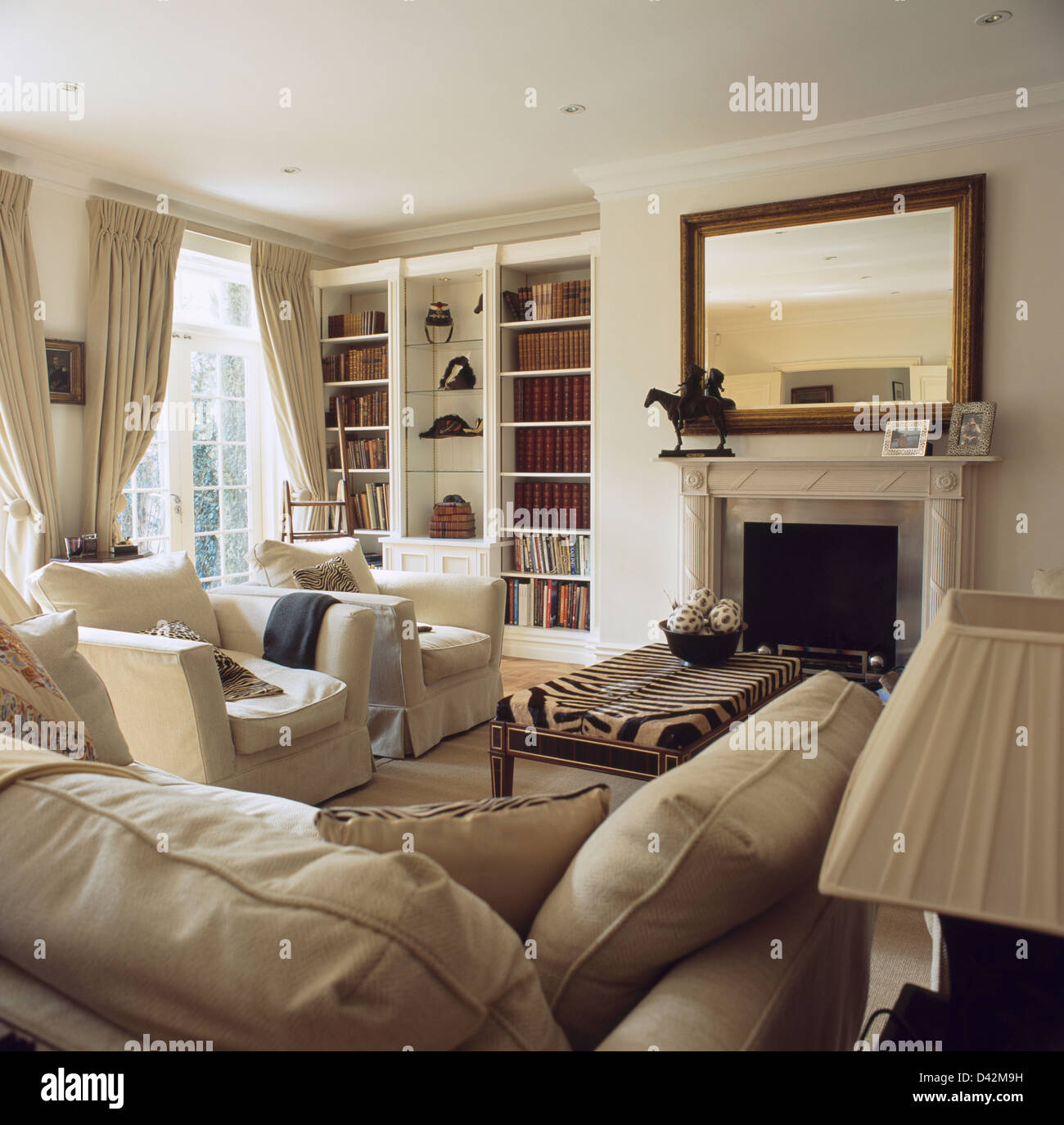 Cremefarbene Couch und Sessel in elegantes Wohnzimmer mit eingebauten  Bücherregal neben Kamin unter großer Spiegel Stockfotografie - Alamy