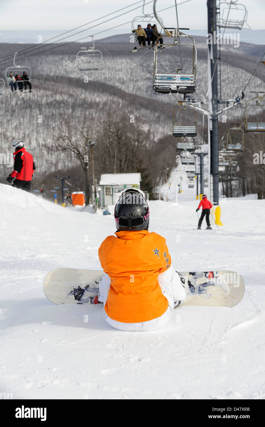 Frau mit Snowboard sitzen und warten am oberen Rand einer Skipiste mit Skilift und Gebirgshintergrund. Stockfoto