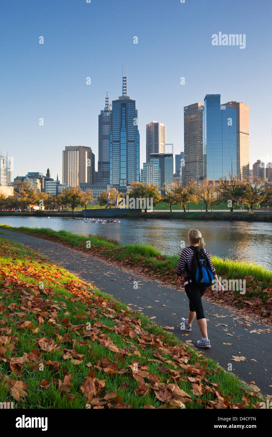 Frau zu Fuß am Ufer des Yarra River mit Skyline der Stadt im Hintergrund. Melbourne, Victoria, Australien Stockfoto