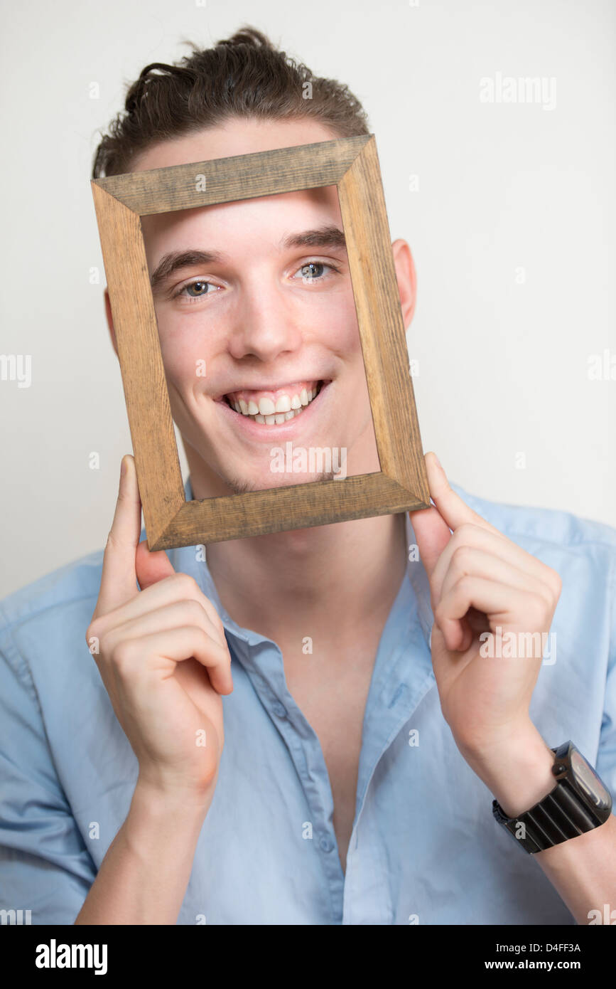 Glücklich und unbeschwert junger Erwachsener Mann posiert mit Holzrahmen vor seinem Gesicht Stockfoto