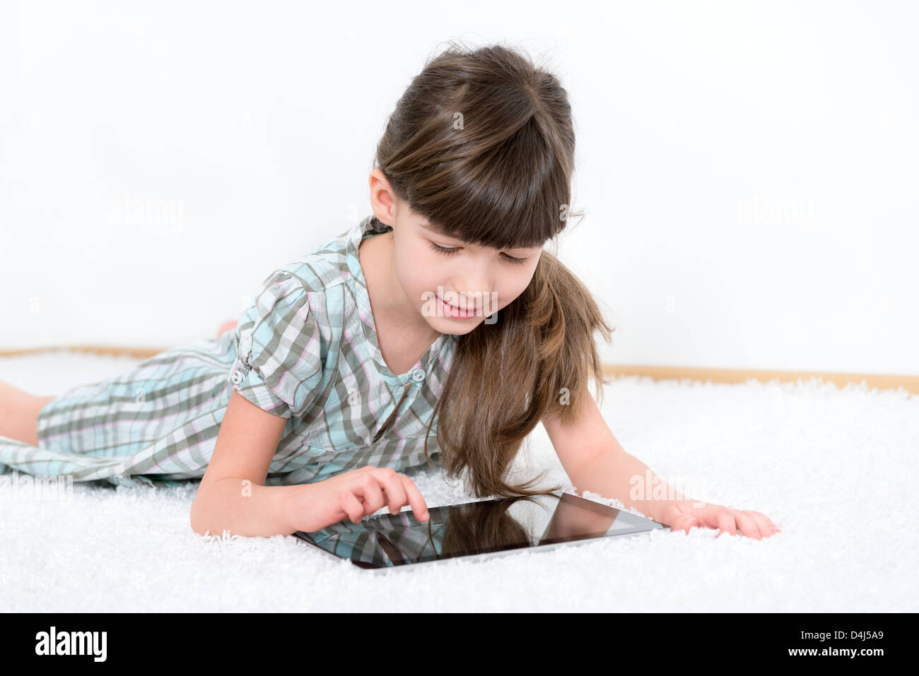 Kleine Mädchen spielen mit einem modernen digitalen Tablet liegend auf einem weißen Teppich in einem weißen Raum Stockfoto