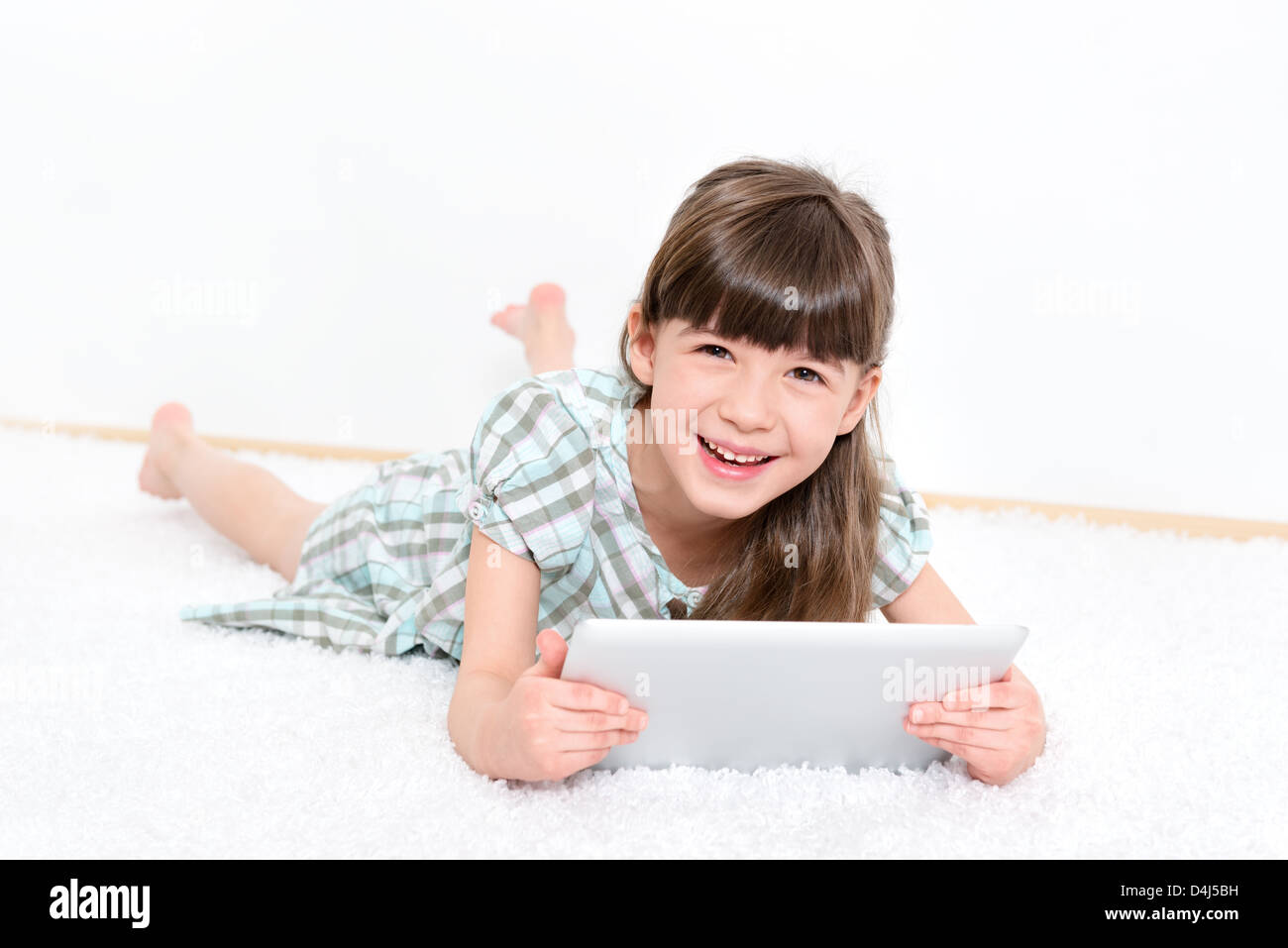 Freudige hübsches kleines Mädchen Lachen als She liegt auf dem Bauch auf einem weißen Teppich mit einem modernen digital-Tablette in einem Raum zu spielen. Stockfoto