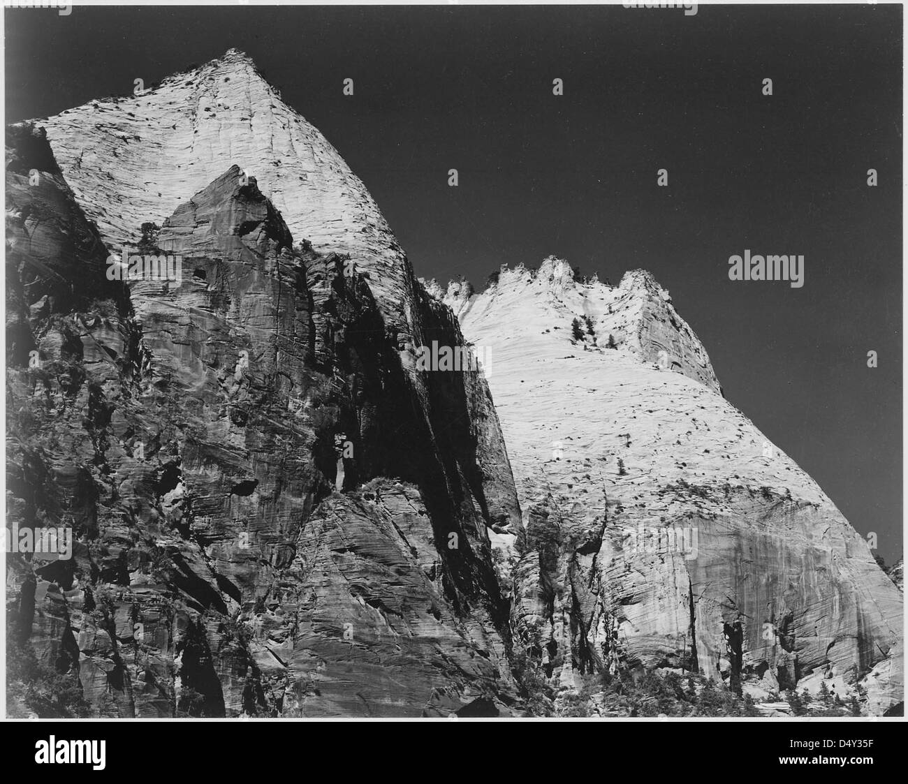 Ein Schwarzweiß-Foto einer Felsformation im Zion-Nationalpark, Utah, aufgenommen 1941 von Ansel Adams. Das Bild fängt die krasse Schönheit der Wüstenlandschaft mit hoch aufragenden Felsformationen vor einem dunklen Himmel ein. Stockfoto