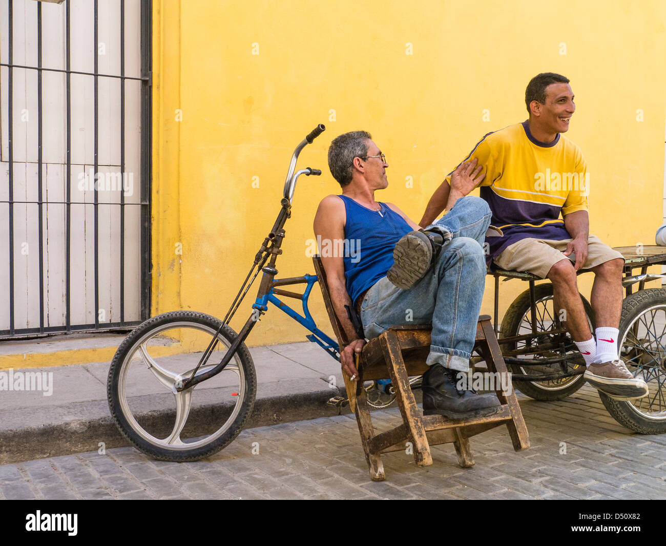 Zwei kubanische Männer sitzen vor einem hellen gelben Wand durch ihre Fahrräder gestikulieren und einem beheizten Gespräch in Havanna, Kuba Stockfoto