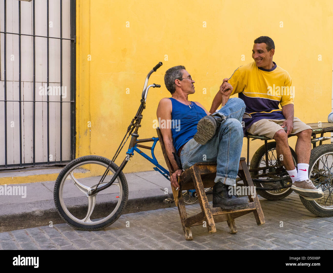 Zwei kubanische Männer sitzen vor einem hellen gelben Wand durch ihre Fahrräder gestikulieren und einem beheizten Gespräch in Havanna, Kuba Stockfoto