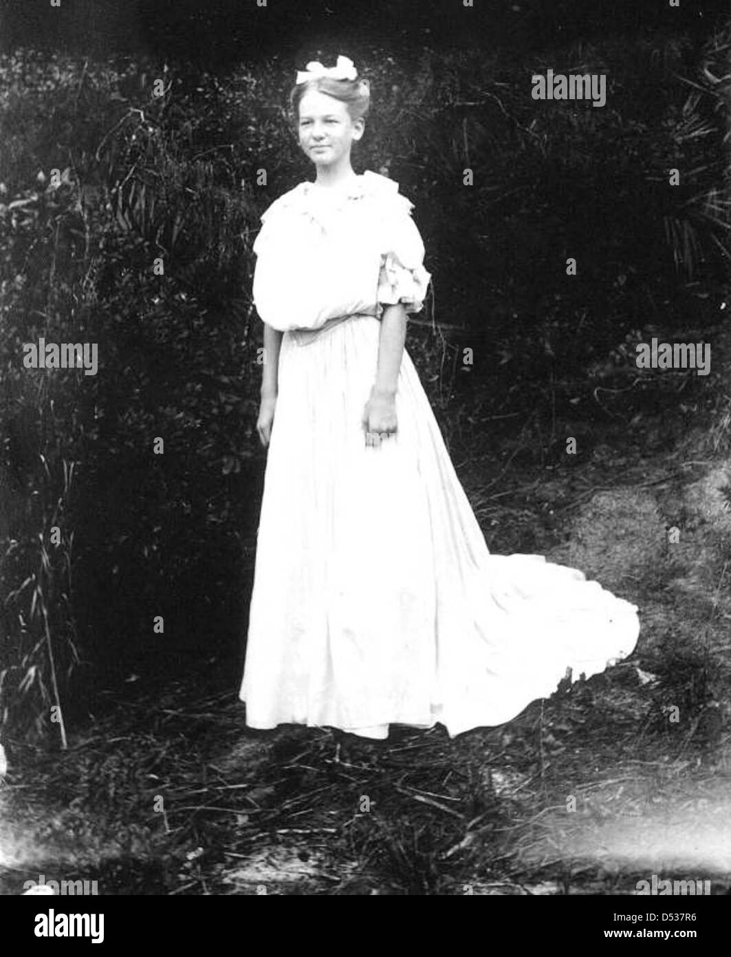 Eine junge Dame trägt ein Kleid mit einem eleganten Zug, wahrscheinlich aus dem frühen 20. Jahrhundert. Das Foto zeigt die Modetrends der Zeit und zeigt das komplizierte Design und die stilvolle Silhouette des Kleides, typisch für diese Zeit. Stockfoto