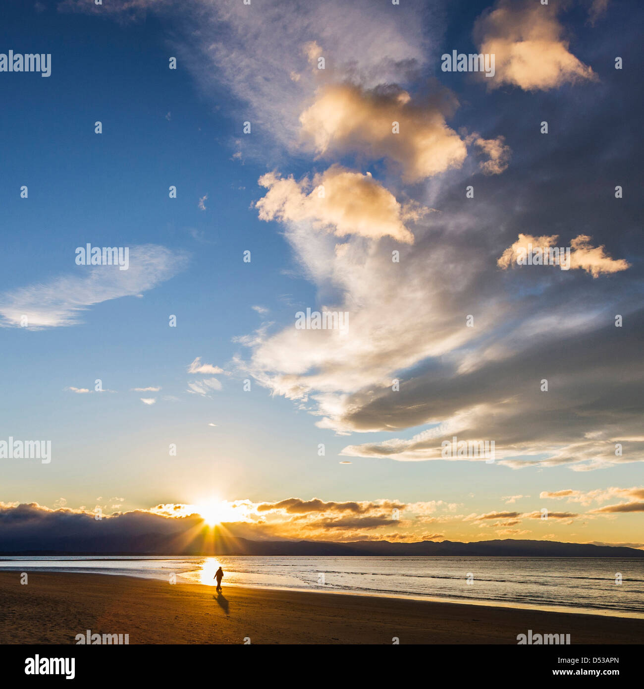 Tahuna Beach, Nelson Bay, Neuseeland. Person zu Fuß am Strand am Rande des Meeres, so wie die Sonne hinter den Bergen verschwindet. Stockfoto