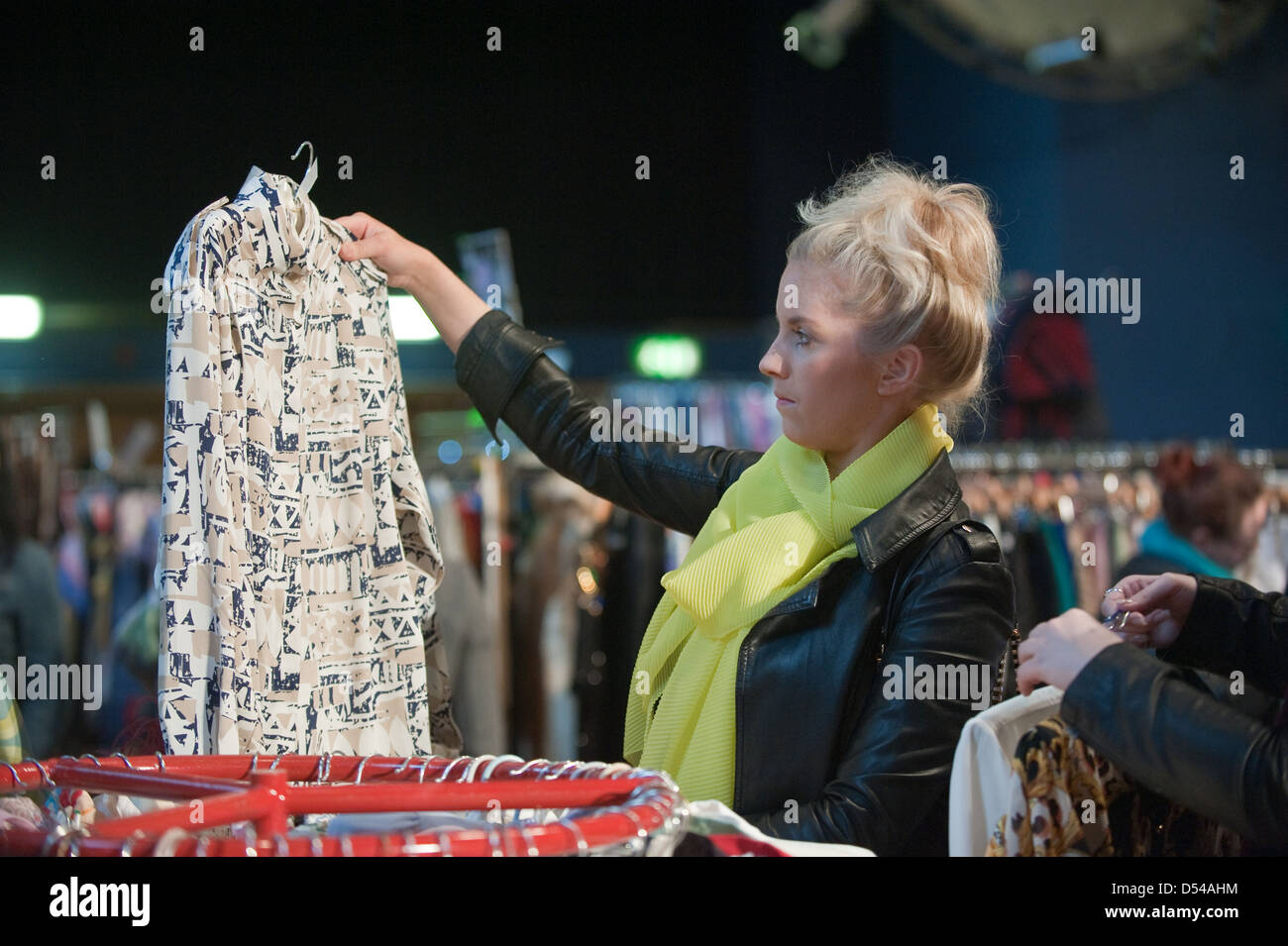 Blonde junge Frau steht unter Kleiderstangen und hält ein Vintage Hemd auf einem Kleiderbügel in einer Vintage-Kleidung fair Stockfoto