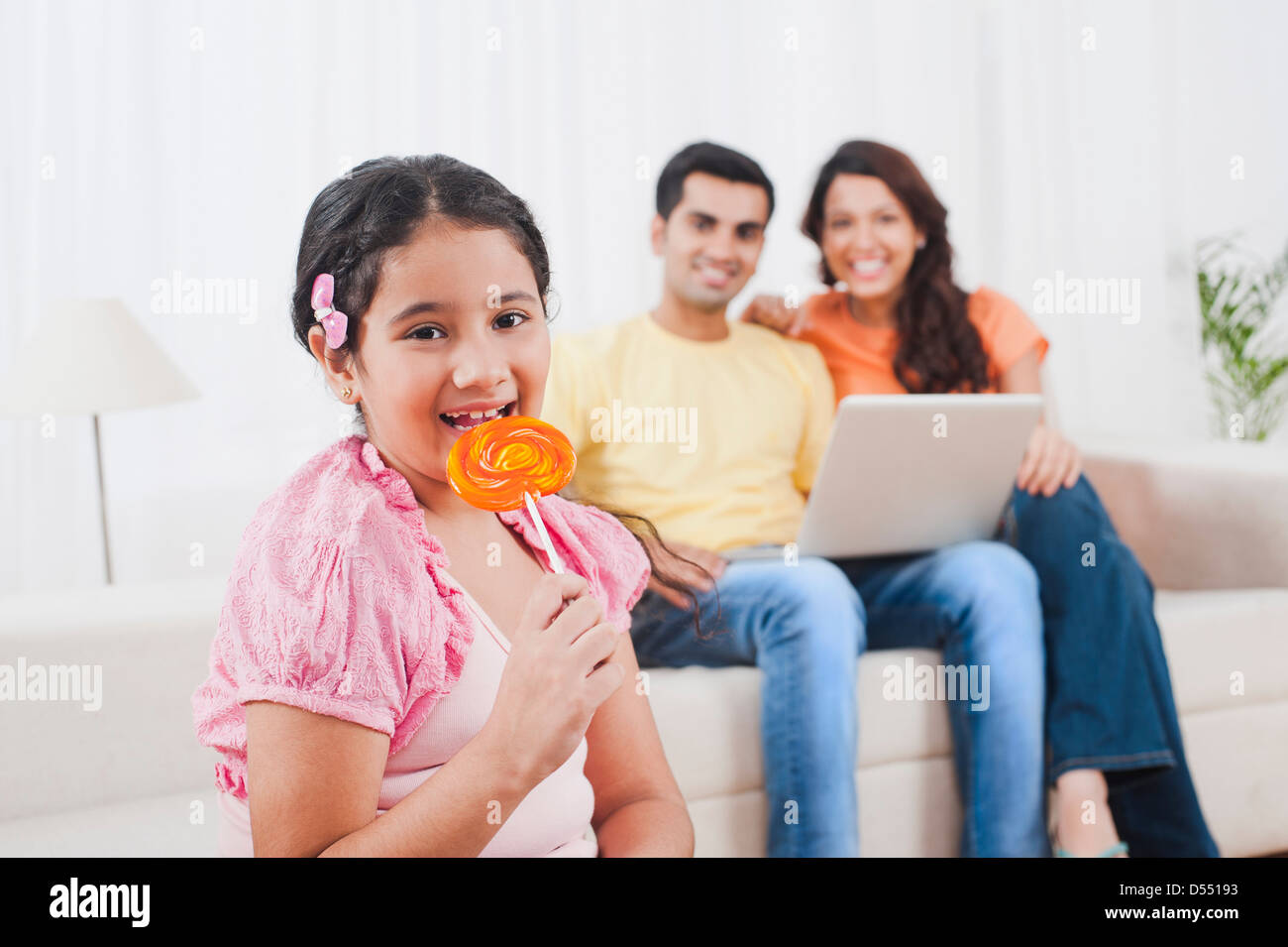 Mädchen essen einen Lolli mit ihren Eltern mit Laptop im Hintergrund Stockfoto