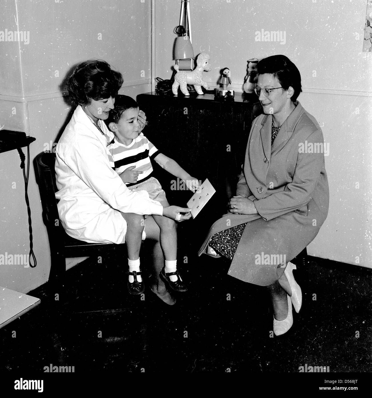 Historische, 1950er Jahre. An einem lokalen Gesundheit Klinik für ein Auge prüfen. Ein kleiner Junge sitzt auf einem weiblichen GP oder Doctor's Lap gezeigt wird, eine Karte zu lesen, mit seiner Mutter Standortwahl neben in unterstützen. Stockfoto