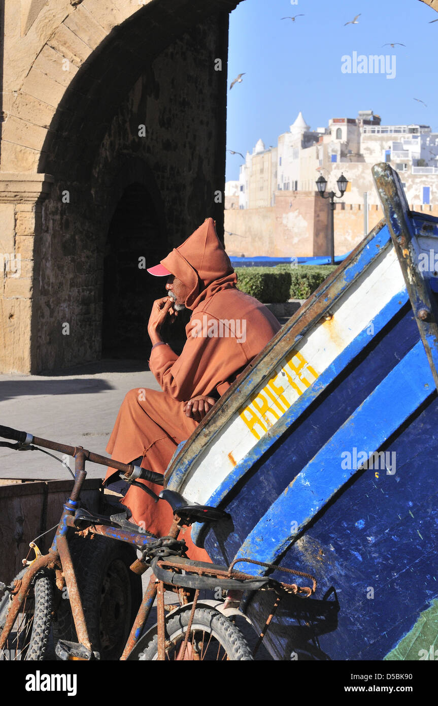 Marokkanischer Mann trägt eine Djellaba die traditionellen Winter Poncho Art Mäntel mit Kapuze, sitzt neben Boot in der Nähe Bab El Marsa Tor Essaouira, Marokko Stockfoto