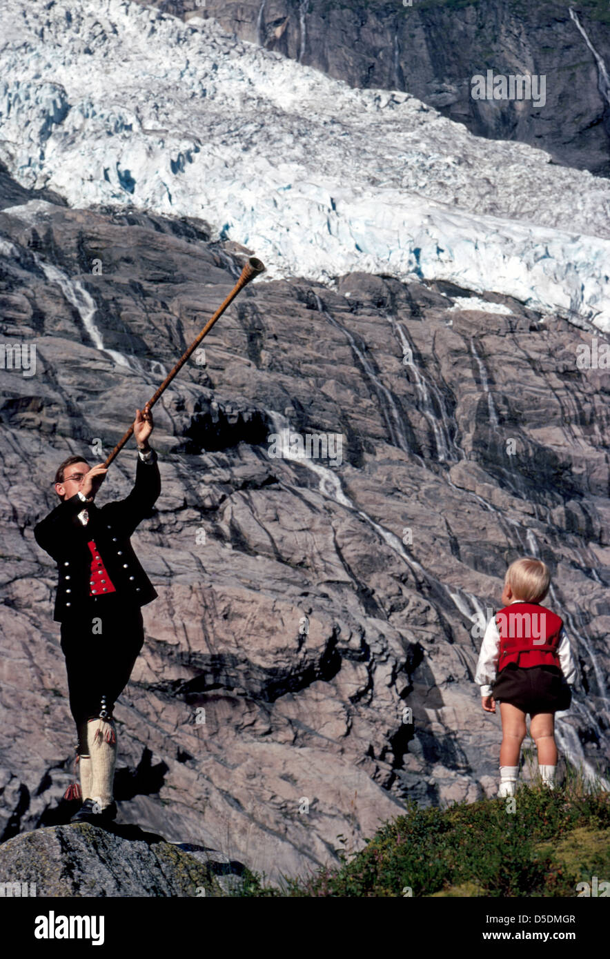 Ein junger Norweger Mann in traditioneller Kleidung vergangener Tage weht ein Vintage Holz Alphorn für seinen Sohn unter einem Gletscher in Fjærland, Norwegen. Stockfoto